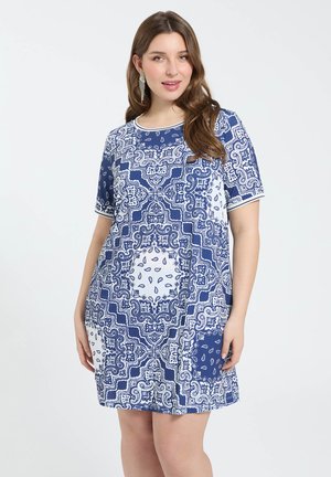 Frau mit langen braunen Haaren, die ein blau-weißes paisley-gestreiftes, kurzärmliges Kleid trägt und vor einem hellgrauen Hintergrund posiert.