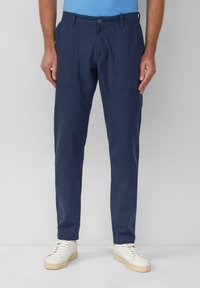 Marineblauwe katoenen broek met een taps toelopende pasvorm, voorzien van zijzakken en een knoopsluiting. Gepaard met witte sneakers op een neutrale achtergrond.