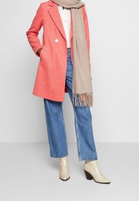 Cappotto in misto lana corallo con design su misura e chiusura a doppio bottone, abbinato a una sciarpa beige con frange, jeans blu a vita alta e stivali beige.