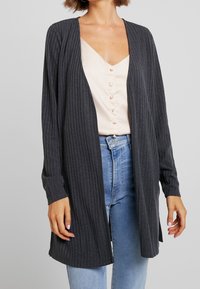 Mörkgrå ribbad cardigan med långa ärmar, öppen framsida och texturerad yta. Buren över en ljusrosa topp och blå jeans.