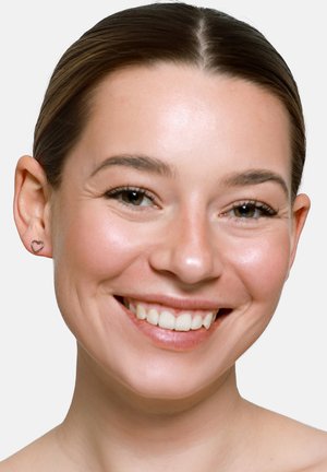 Junge Frau mit hellem Teint und zurückgebundenem braunem Haar, lächelnd, trägt kleine herzförmige Ohrringe, vor einem einfarbigen hellen Hintergrund.