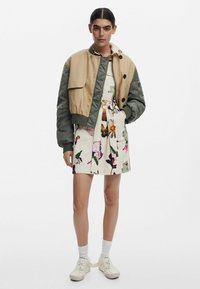 Lichtgewicht bomberjack in bruin en groen, over een jurk met bloemmotief. Sneakers met witte straps, enkelsokken en zichtbare bloemmotieven.