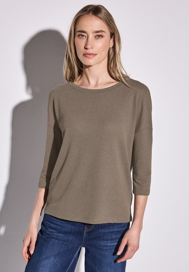 Lehký, texturovaný olivově zelený top s třičtvrtinovými rukávy a kulatým výstřihem, v kombinaci s tmavě modrými džíny.