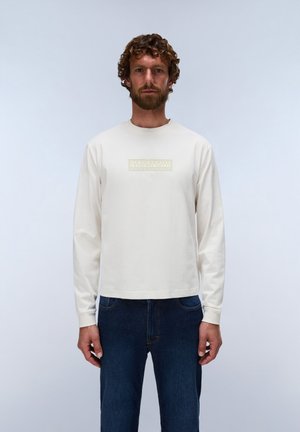 Uomo con capelli ricci e barba che indossa una maglia bianca a maniche lunghe con un piccolo logo sul petto e jeans blu scuro, in piedi davanti a uno sfondo semplice.