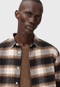 Rutigt skåp med ett mönster i svart, brunt och kräm; flanellmaterial; knäppning fram; parat med en strukturerad brun stickad tröja under.