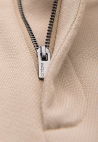 Gros plan d'un curseur de fermeture éclair argenté sur un tissu beige texturé, montrant le nom de la marque « ombre » sur la tirette.