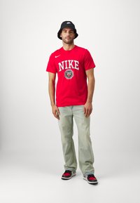 Punane Nike T-särk valge logo ja graafikaga, helesinised teksased ja must ämbri müts, kombinatsioonis mustade ja punaste tossudega.