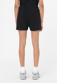 Pantalones cortos de algodón negros con una cintura elástica y un bolsillo trasero, con un corte relajado y una textura suave. Llevados con zapatillas grises.