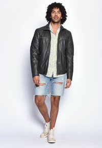 Schwarze Lederjacke mit gesteppten Schulterdetails, getragen über einem floralen Hemd, kombiniert mit zerrissenen blauen Denim-Shorts und weißen Sneakers.