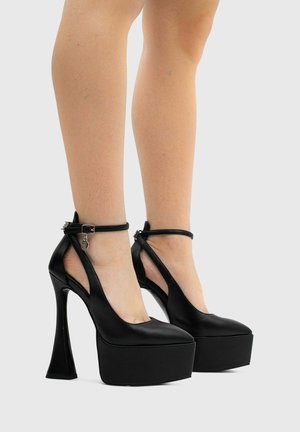 Gambe con tacchi alti platform neri con cinturini alla caviglia e un piccolo charm argentato su uno sfondo chiaro e uniforme.