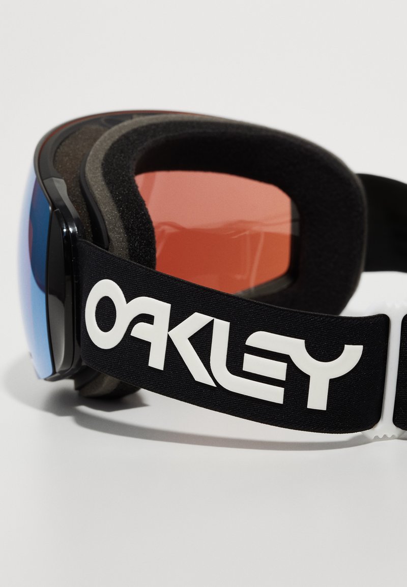 Gafas de esquí negras con lentes reflectantes en azul y rojo, marco de espuma acolchada y una correa negra con la marca "OAKLEY" en blanco.