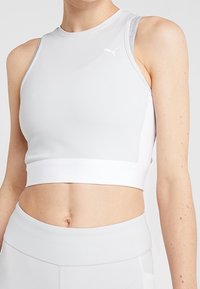 Top deportivo corto gris claro hecho de un material suave y elástico, con un cuello alto y una cinturilla blanca en contraste. Logo de Puma en la parte frontal.
