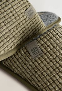 Teksturowane oliwkowe klapki z pluszowym wierzchem i kontrastującym szarym wykończeniem, z delikatną łatką z logo z boku.