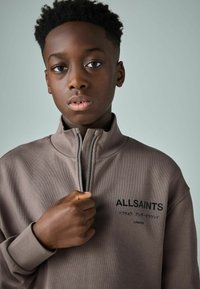 Felpa marrone con zip e polsini a costine, con il logo "ALLSAINTS" in nero, indossata da un giovane, su uno sfondo chiaro.