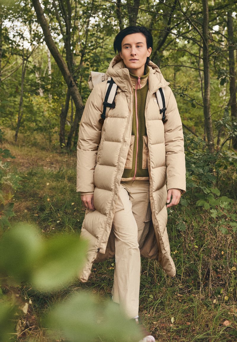 Piumino lungo beige con cappuccio, caratterizzato da ampie sezioni trapuntate. Indossato sopra una felpa verde con zip, abbinato a pantaloni beige e uno zaino.