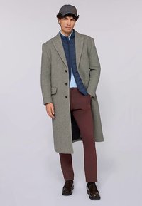 Grå yllekappa med strukturerade slag, mörkblå quiltad väst, ljusblå skjorta, burgundyfärgade byxor och mörka skor. Casual keps ingår.