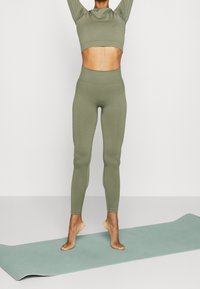 Olivgrön sportcrop-top och matchande leggings, tillverkade av ett följsamt tyg med en figurnära design, står på en mintgrön yogamatta.
