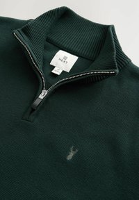 Sudadera verde oscuro con cierre completo, cuello acanalado, tejido texturizado y pequeño logo de ciervo. Cuenta con una cremallera completa y etiqueta de talla.