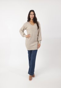 Pull en tricot beige avec un col en V et des manches longues légèrement froncées, associé à un jean bleu foncé et des sandales transparentes.