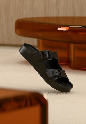 Black slip-on sandal with two adjustable buckles floating above a light-colored carpet in a room with amber-tinted furniture.