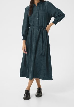 Woman wearing a dark teal long-sleeve midi dress with button front, tied waist belt, and black chunky loafers standing on white background.