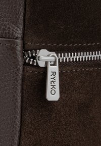 Närbild av en silver dragkedjehandtag graverad med "RYŁKO" på mörkbrun mocka- och lädertyg.