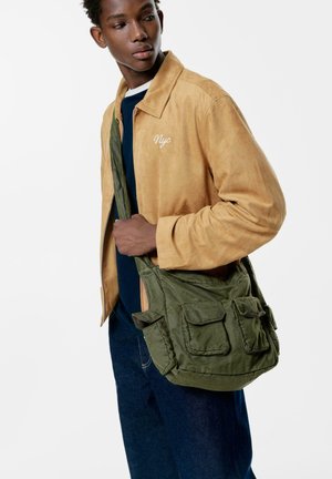 Veste en daim tan avec un détail brodé "NYC", portée sur une chemise bleue. Un sac en toile verte avec plusieurs poches est porté en travers du corps.