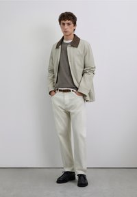 Ljusbeige jacka med brun krage, grå tröja under, cremefärgade byxor och svarta skor. Enkel design med en detalj i form av en framficka.