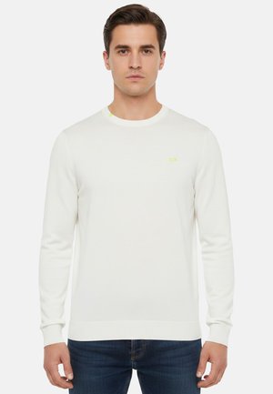 Uomo che indossa un maglione bianco semplice a maniche lunghe e girocollo e jeans blu scuro, in piedi davanti a uno sfondo chiaro e uniforme.