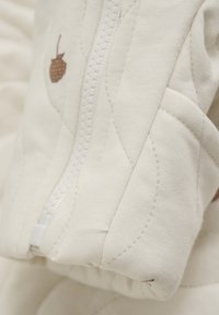 Tissu matelassé beige clair avec un détail de fermeture éclair. Présente un petit motif de gland marron et des coutures visibles. Texture douce et finition lisse.