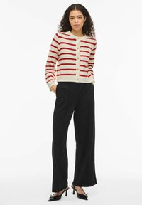 Cardigan rayé rouge et blanc avec une fermeture à boutons, poignets et ourlet côtelés, associé à un pantalon large noir et des chaussures à talons noires.