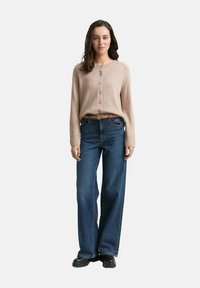Frau steht, trägt eine beigefarbene Knopfjacke, die in hochtaillierte, weite blaue Jeans mit einem braunen Gürtel und schwarzen Schuhen gesteckt ist.