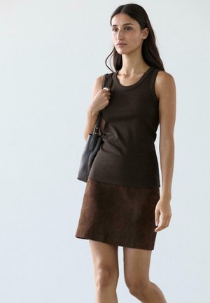 Donna che indossa una maglietta scura senza maniche e una gonna marrone, che tiene una borsa nera a tracolla, in piedi davanti a uno sfondo chiaro uniforme.