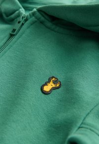 Sweat à fermeture éclair vert en tissu doux, avec un logo de homard brodé jaune sur un contour noir. Texture lisse, coupe standard.