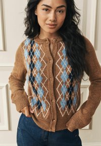 Kvinna iförd en brun knappad kofta med blå och vit argyle-mönster, händerna i fickorna, och mörkblå jeans.