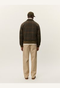 Chaqueta bomber de cuadros marrones con puños y dobladillo de canalé, llevada sobre una camisa blanca. Pantalones beiges completan el conjunto, vistos desde atrás.