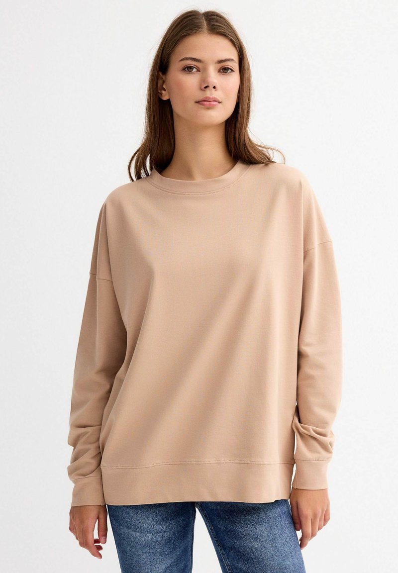 Sudadera beige con cuello redondo, hombros caídos y mangas largas. Textura suave y corte relajado. Combinada con vaqueros azules.
