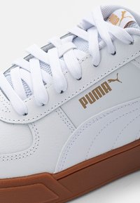 Zapatilla Puma blanca con cordones blancos, logo de Puma dorado y suela de goma marrón, mostrando costuras detalladas y cuero texturizado.