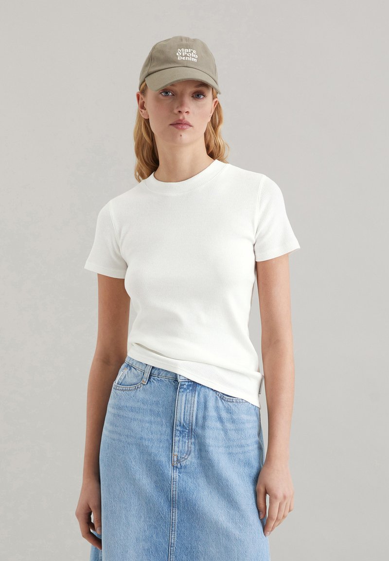Weißes, geripptes Kurzarmoberteil, kombiniert mit einem hellblauen Jeansrock. Eine hellolivfarbene Kappe mit weißem Text sitzt auf dem Kopf des Models.