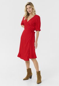 Robe rouge avec des manches courtes à volants, un ourlet froncé et une taille à nouer. Portée avec des bottes chevilles en daim marron avec un talon bloc.