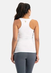Weißes ärmelloses Tanktop mit taillierter Passform und rundem Ausschnitt. Mit einem kleinen goldenen Logo auf der Rückseite. Kombiniert mit grauen Leggings.