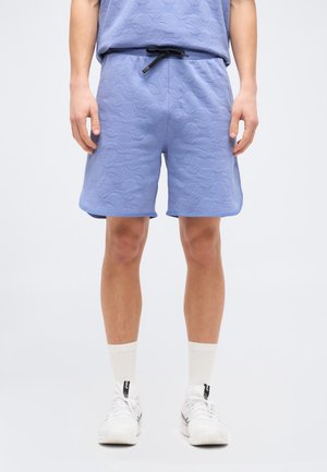 Άτομο που φοράει ανοιχτό μπλε υφασμάτινο σορτς με μαύρο κορδόνι, λευκές κάλτσες μέχρι τον αστράγαλο και λευκά αθλητικά παπούτσια, στέκεται σε απλό φόντο.