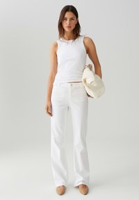 Top bianco senza maniche e jeans bianchi a palazzo, abbinati a ballerine beige, mentre tiene una borsa chiara. Design semplice con una texture liscia.