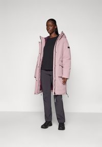 Roze geïsoleerde parka met een capuchon, twee voorzakken en zwarte ritsdetails. Draagt het over een zwart shirt en gecombineerd met grijze broek en zwarte schoenen.