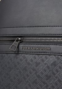 Black synthetic backpack featuring a horizontal zippered pocket, embossed pattern, and a metal logo plate showcasing "TOMMY HILFIGER."