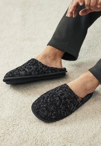 Chaussons tricotés noirs avec un motif texturé et une semelle rembourrée, ornés d'une étiquette en cuir marron. Portés pieds nus, sur un tapis clair.