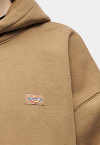 Ύφασμα καφέ φούτερ με μικρό ορθογώνιο ετικέτα της μάρκας Dickies ραμμένη στην περιοχή του στήθους, εμφανίζοντας τμήμα της κουκούλας και της ραφής του μανικιού.