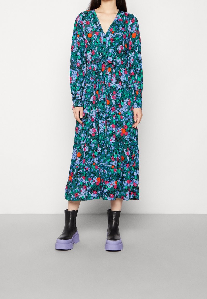 Femme portant une robe midi verte à manches longues avec un motif floral et une ceinture à la taille, associée à des bottines noires avec des semelles épaisses violettes.