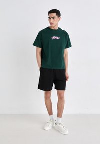 Camiseta de algodón verde con un logo gráfico rosa en el pecho, combinada con pantalones cortos negros y zapatillas blancas. Diseño simple, corte relajado.