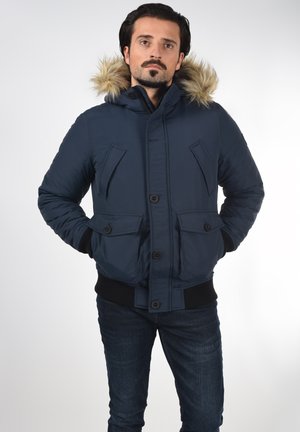 Navy puffer jacket with a fur-lined hood, featuring four front pockets, a button closure, and ribbed cuffs for a snug fit.